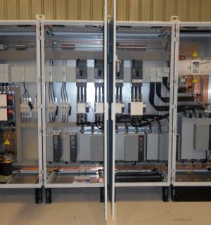 Three open electrical control cabinets with wiring, circuit breakers, and motor drives in a machinery room.