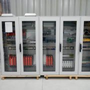 Four gray electrical control cabinets with glass doors on a wooden pallet in a warehouse; internal wiring and red components visible behind glass.