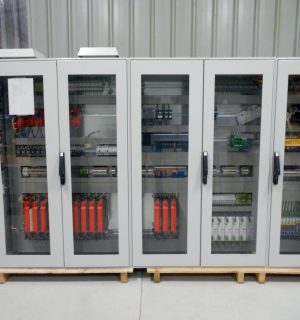 Four gray electrical control cabinets with glass doors on a wooden pallet in a warehouse; internal wiring and red components visible behind glass.