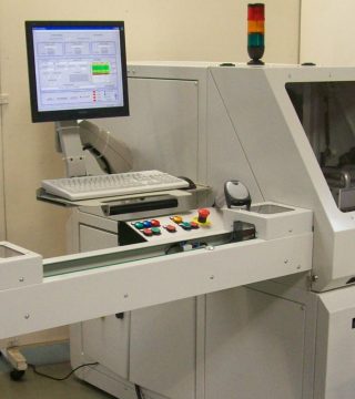 Industrial manufacturing machine with computer control monitor, keyboard, and a row of colored control buttons on a conveyor belt.